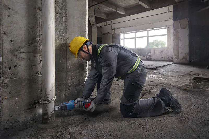 Güvenlik ekipmanı takan bir kişi, beton bir duvarın yakınındaki bir boruyu kesmek için alternatif hareketli bir testere kullanıyor.