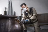 Güvenlik ekipmanı takan bir kişi büyük bir metal borunun kenarını taşlıyor.