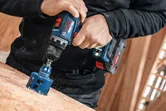 Bir kişi atölyede elektrikli alet kullanarak tahtaya büyük bir delik açıyor.