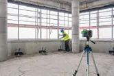Güvenlik ekipmanı giyen bir kişi, üç ayaklı bir sehpa üzerindeki lazerli seviyeleme aletini kullanarak duvarı hizalıyor.