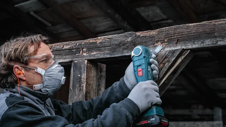 Ahşap bir kirişi zımparalamak için kablosuz bir alet kullanan güvenlik ekipmanı takan kişi.