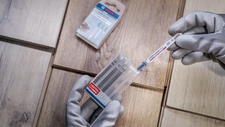 Güvenlik ekipmanı takan kişi, ahşap fayansların üzerindeki plastik bir kutudan bir testere bıçağı seçiyor.