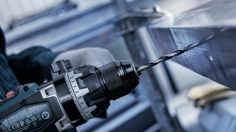 Güvenlik ekipmanı takan bir kişi, elektrikli matkapla metal bir kirişe delik açıyor.