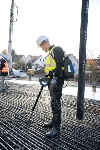 Güvenlik ekipmanı giyen bir kişi, demir donatı ızgarası üzerinde beton vibratörü kullanıyor.