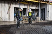Güvenlik ekipmanları giyen iki işçi, demir donatılı bir levha üzerindeki betonu sıkıştırmak için elektrikli aletler kullanıyor.