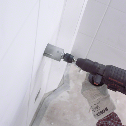 Worker wearing safety equipment drills a large circular hole into tiled wall.