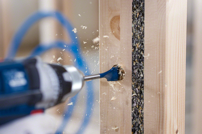 Person wearing safety equipment drills a hole in a wooden frame.