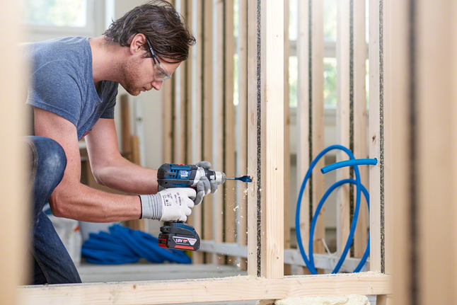 A worker wearing safety equipment drills a hole through a wooden stud.