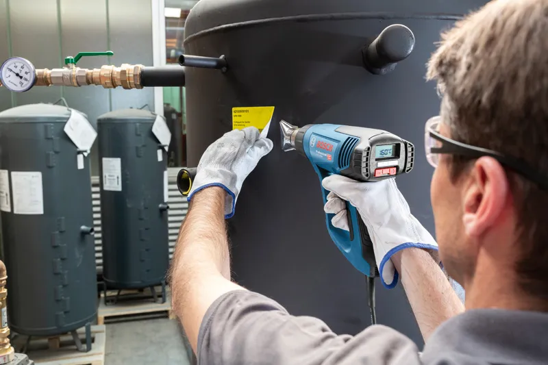 A person wearing safety equipment uses a heat gun to apply a label to a large tank.