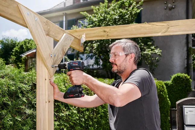 Person wearing safety equipment drills screws into a wooden beam outdoors.