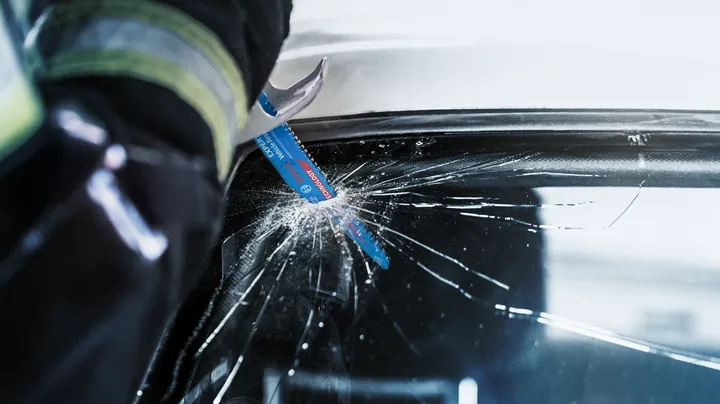 Person wearing safety equipment cuts a cracked car windshield with a reciprocating saw blade.
