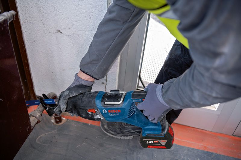 A person wearing safety equipment uses a reciprocating saw to cut metal near a door frame.
