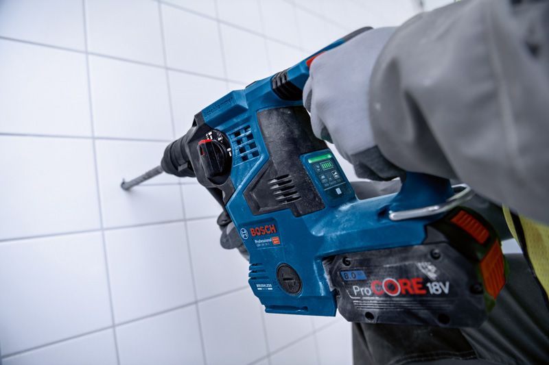 A person wearing safety equipment drills into a tiled wall using a cordless rotary hammer.