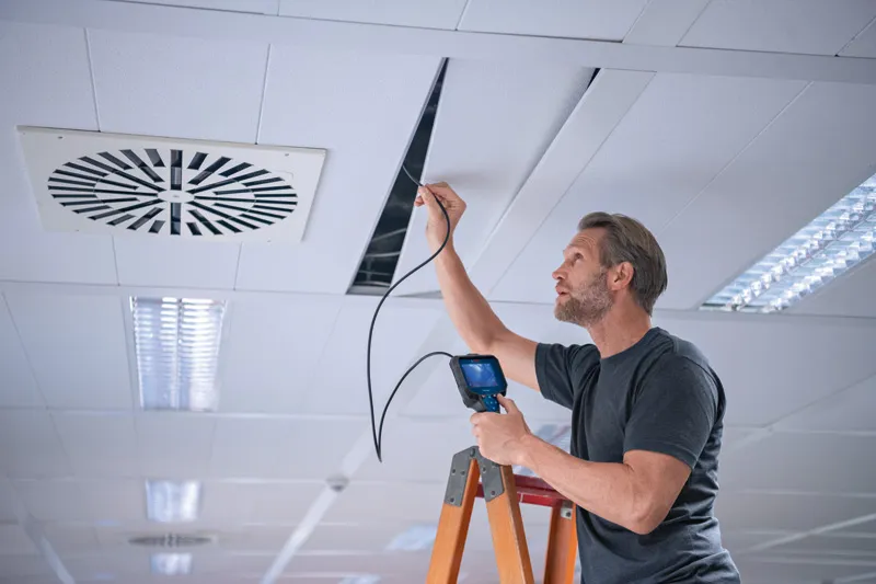 A person on a ladder inspects a ceiling panel with an inspection camera.