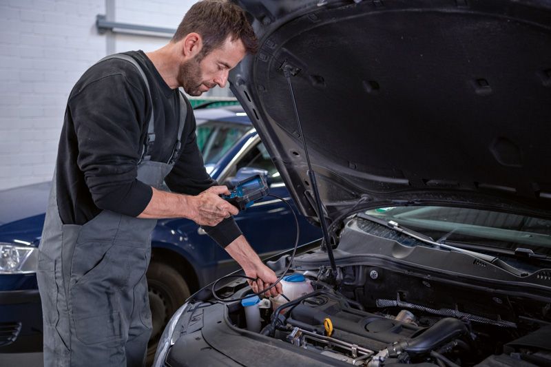 A person in coveralls inspects a car engine bay using an inspection camera.