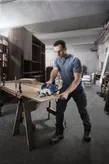 A person wearing safety equipment cuts wood with a cordless circular saw in a workshop.