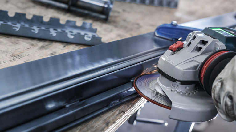 Person wearing safety equipment grinds a metal beam with an angle grinder.