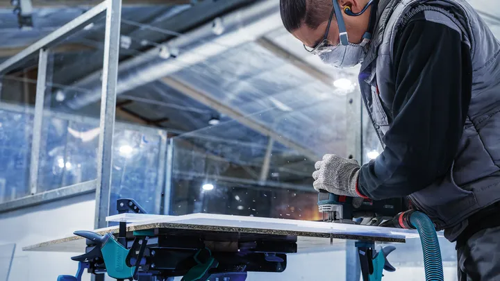 A person wearing safety equipment uses a power tool to cut a wood panel.