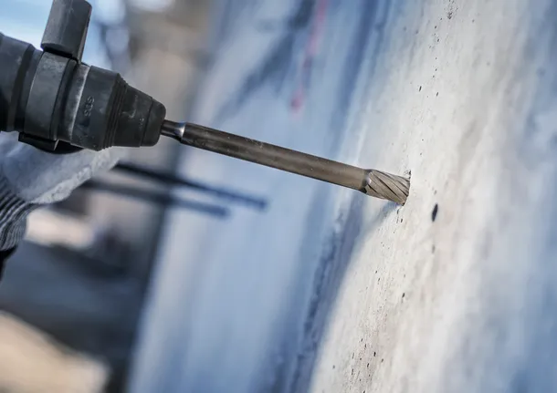Person wearing safety equipment drills into concrete with a rotary hammer.