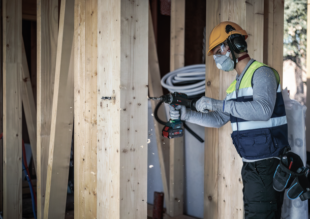 Person wearing safety equipment drills into a wooden beam on a construction site.