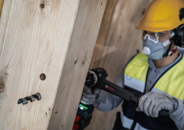 Person wearing safety equipment drills a large hole into thick wooden beams.