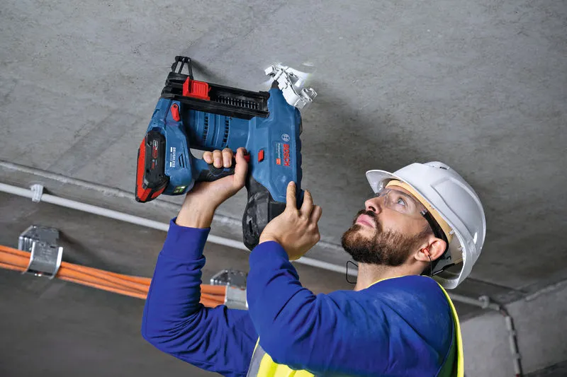 A person wearing safety equipment uses a cordless concrete nailer on a ceiling.