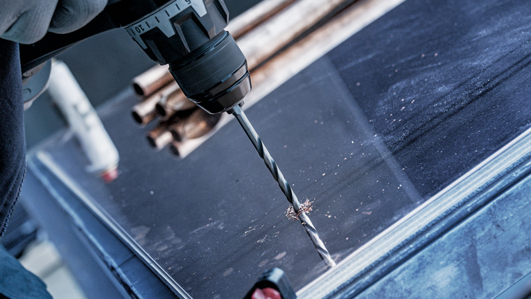 A person wearing safety equipment drills into a metal sheet on a workbench.
