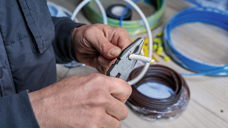 Person uses pliers to strip insulation from electrical wire on a workbench.
