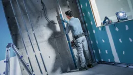 Person wearing safety equipment measuring a wall with construction tools nearby.