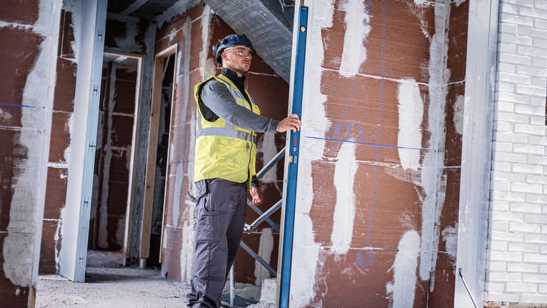 Person wearing safety equipment checks wall alignment with a digital level.