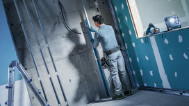 A person wearing safety equipment measures a metal stud frame at a construction site.