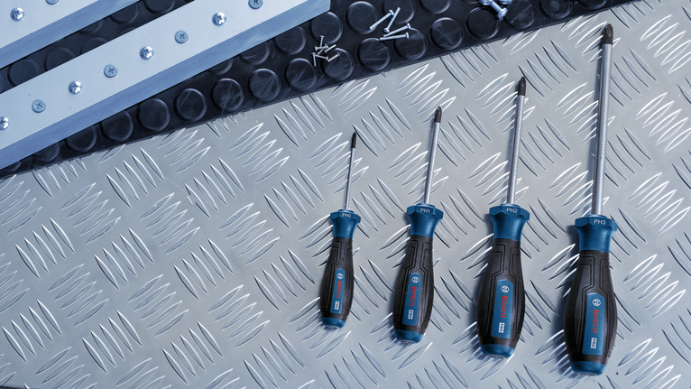 Four screwdrivers arranged on a metal surface beside screws and metal strips.
