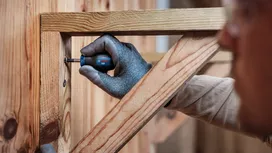 Person wearing safety equipment uses a screwdriver to fasten a screw into wooden beams.