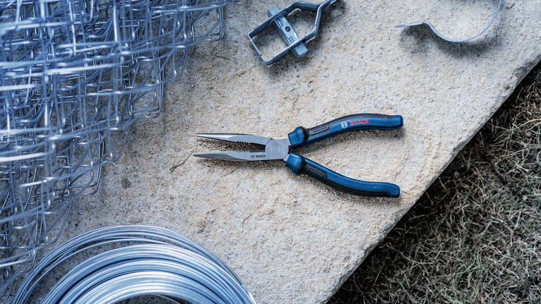 Needle-nose pliers placed on a stone surface next to wire mesh and metal wires.