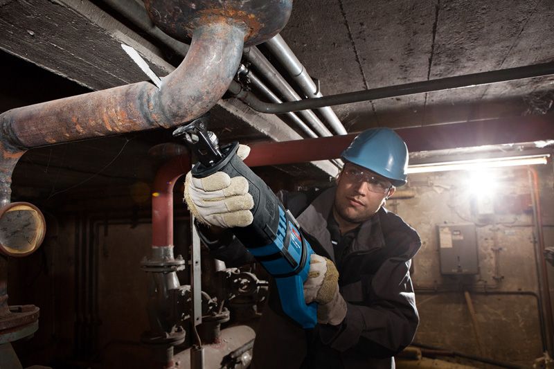 A person wearing safety equipment cuts a metal pipe with a reciprocating saw.