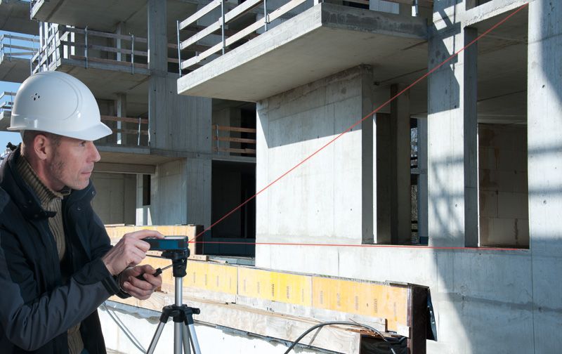 A person wearing safety equipment uses a laser measure on a tripod at a construction site.