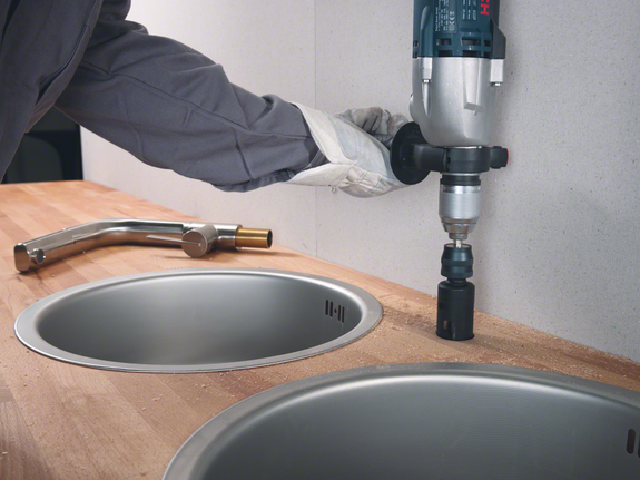 Person wearing safety equipment drills a hole in a countertop beside a kitchen sink.