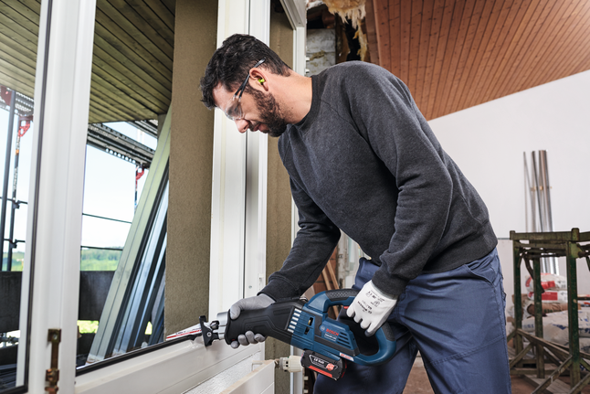 Працівник у захисному спорядженні використовує зворотно-поступальну пилку для різання біля віконної рами.