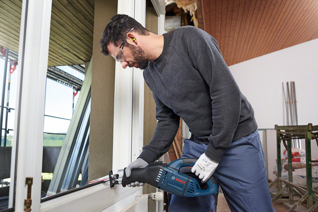 Людина в захисному спорядженні використовує зворотно-поступальну пилку для розрізання віконної рами.