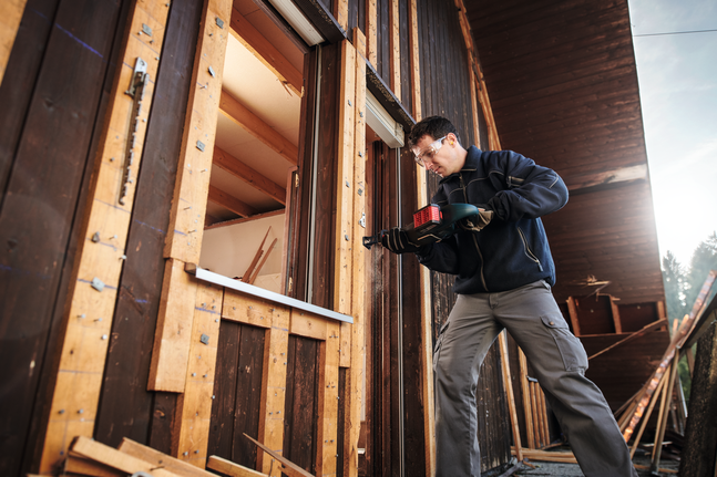 Працівник у захисному спорядженні використовує акумуляторний дриль на дерев'яній віконній рамі.