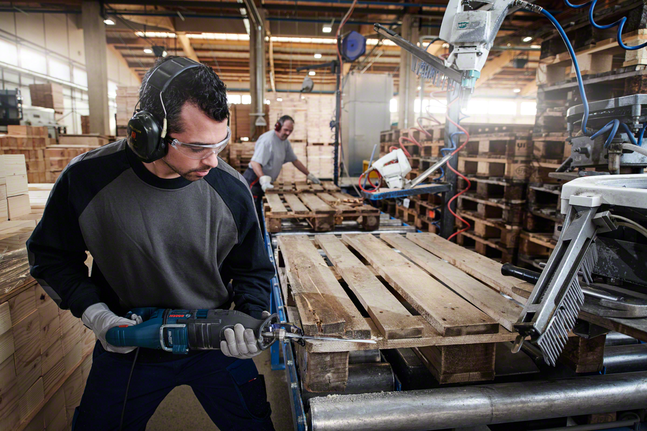 Людина в захисному спорядженні використовує зворотно-поступальну пилку для розпилювання дерев'яного піддону.