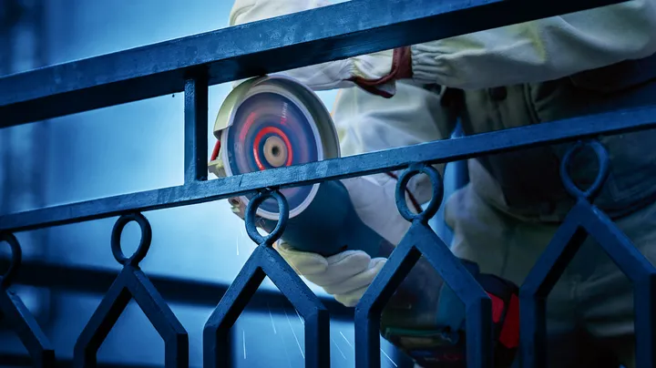 Людина в захисному спорядженні шліфує металевий паркан електроінструментом.