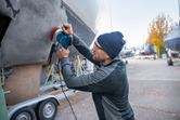 Людина в захисному спорядженні шліфує корпус човна на відкритому повітрі за допомогою ексцентрикової шліфувальної машини.