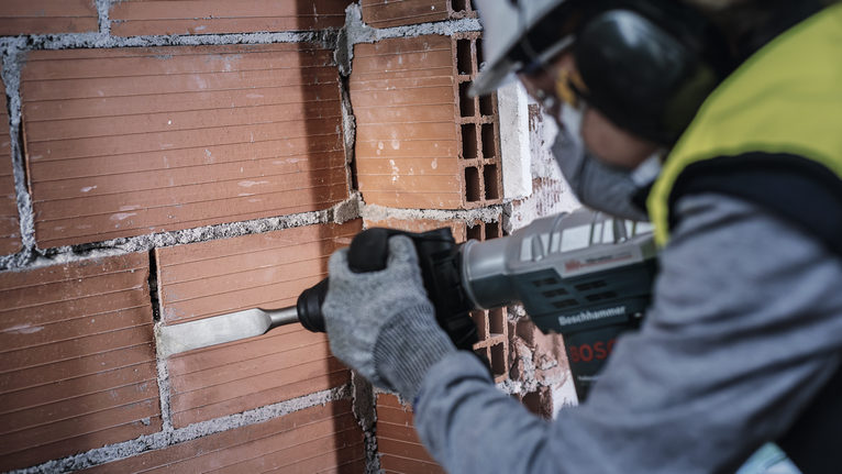 Людина в запобіжному спорядженні долобить цегляну стіну за допомогою електроінструменту.