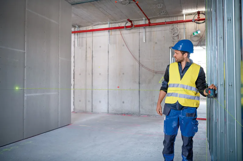 Una persona que usa equipo de seguridad utiliza una herramienta niveladora láser para alinear la estructura de la pared.
