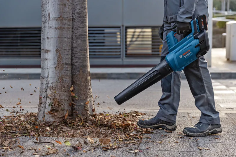 Una persona que lleva equipo de seguridad utiliza un soplador de hojas inalámbrico para mover las hojas cerca de un árbol.