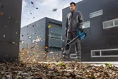Una persona con equipo de seguridad utiliza un soplador de hojas inalámbrico para limpiar las hojas cerca de edificios modernos.