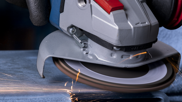 Una persona que usa equipo de seguridad muele metal con una amoladora angular, creando chispas.