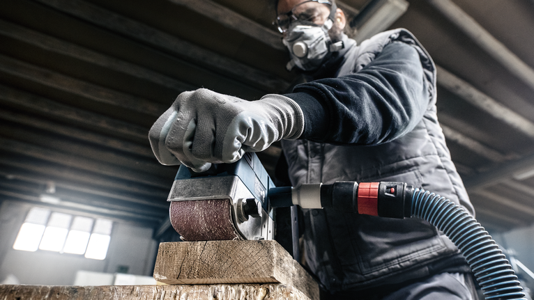 Persona con equipo de seguridad lijando una viga de madera utilizando una herramienta eléctrica.