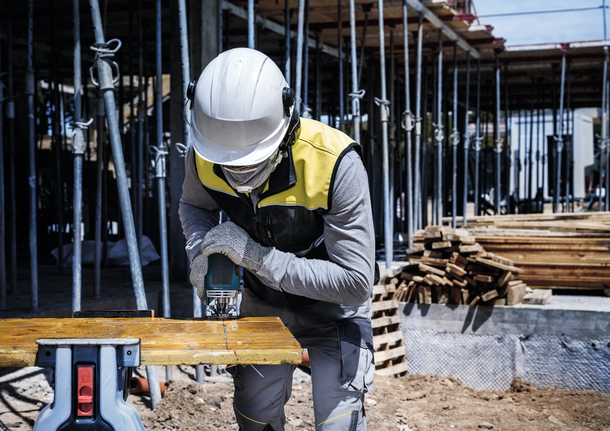 Una persona que usa equipo de seguridad corta una tabla de madera con una herramienta eléctrica en un sitio de construcción.
